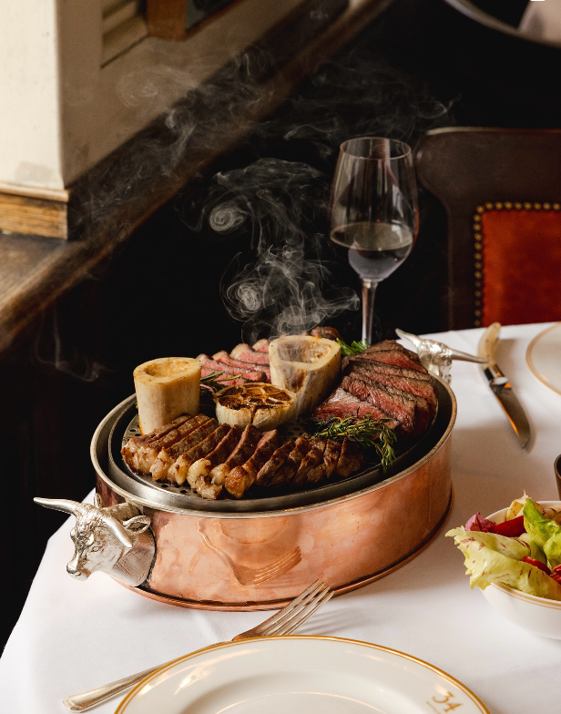 A copper pan of grilled cuts and bone marrow, smoke rising past a glass of red wine